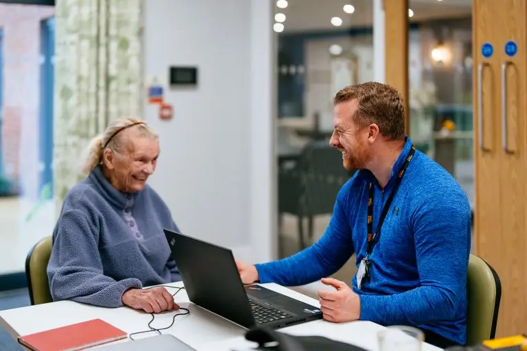 A male member of staff speaking to woman whilst using a laptop.
