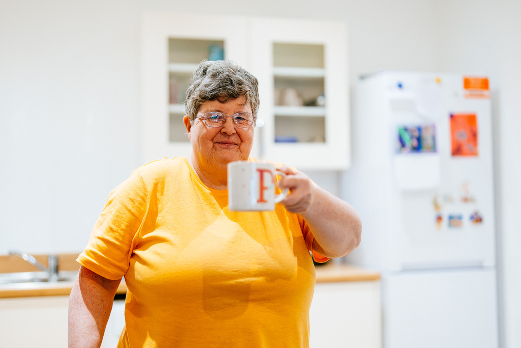 A female resident holding a cup of tea. She is wearing an orange top and is stood in her kitchen.