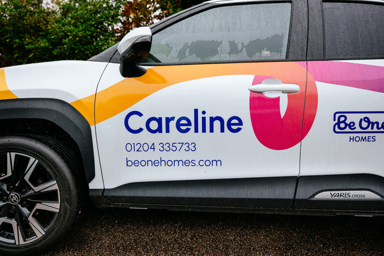 The side of a Be One Homes Careline van. The car has our multicoloured 'brand thread', the Careline logo, and the Be One Homes phone number and web address.
