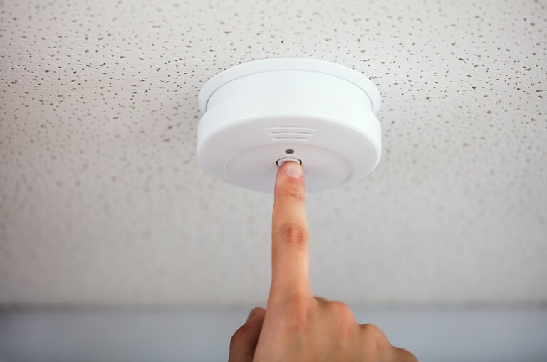 A smoke detector/alarm fixed to a ceiling. Someone is pressing the test button with their index finger.