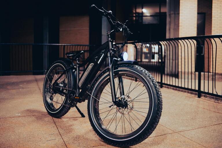 A black electric bike. It is stood upright on its kick stand. There are metal railings in the background.