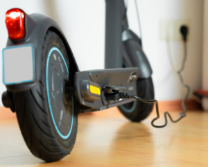 An electric scooter plugged into a wall socket to charge. The wheels are visible and the scooter is placed on a laminate floor.