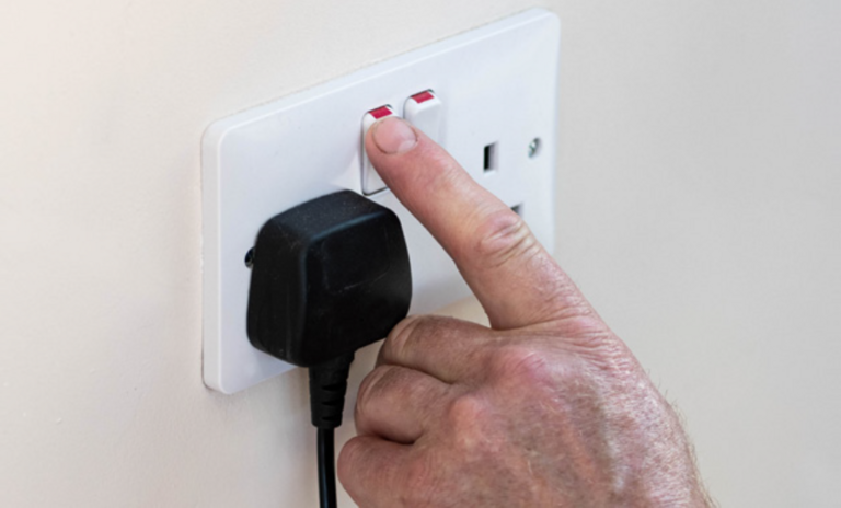 A dual plug wall socket with a black power plug connected to the left socket. Someone is turning off one of the switches with their finger.